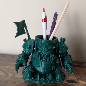 Op de afbeelding: Een donkergroene pennenhouder in de vorm van een fantasiefiguur, met een vlag en een bijl. Het bevat een witte en rode pen, een paarse pen en een houten potlood. De pennenhouder staat op een houten oppervlak.