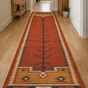 May include: A long, rectangular rug with a rust-coloured field and geometric patterns. The rug features a central tree-like design in black, with borders in shades of gold, navy, and cream. The rug is placed in a hallway.