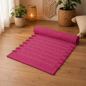 Puede incluir: Una alfombra magenta enrollada con rayas horizontales blancas y flecos. La alfombra está hecha de material tejido y está parcialmente desenrollada sobre un piso de madera. Es un artículo de decoración del hogar.