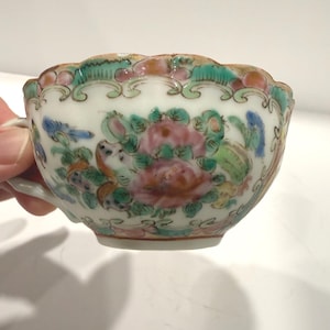 May include: A white porcelain teacup with a floral design in pink, green, blue, and yellow. The cup has a scalloped rim and a small handle. The floral pattern includes peonies and other flowers.