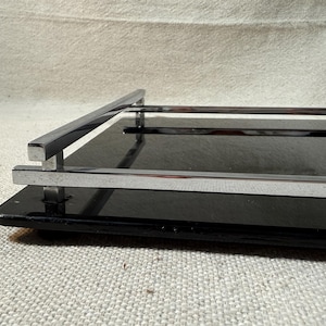 May include: A rectangular black serving tray featuring a polished chrome metal railing along the sides. The tray has a smooth, glossy black surface and is supported by small feet, with the metal frame extending slightly above the base.