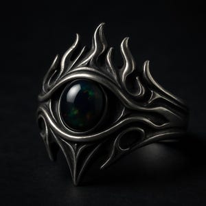 Può includere: Un anello d'argento con un design a forma di occhio. L'anello presenta una gemma scura di forma ovale al centro, circondata da un motivo dettagliato a forma di fiamma. L'anello è posto su uno sfondo scuro, evidenziando il suo design intricato.