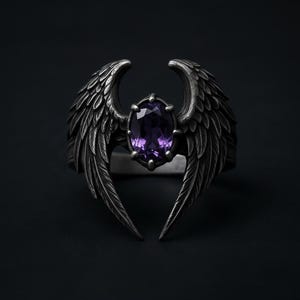 Puede incluir: Anillo plateado con una gema central ovalada de color p&uacute;rpura, flanqueada por dise&ntilde;os de alas detallados. Las alas se curvan hacia arriba, creando una est&eacute;tica &uacute;nica. El anillo se encuentra sobre un fondo oscuro.