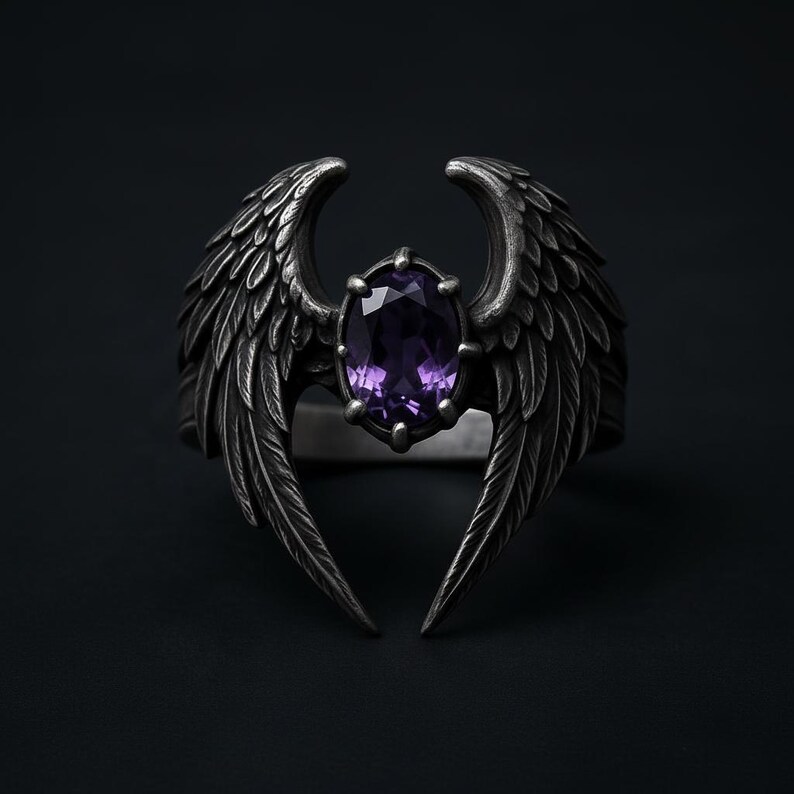 Puede incluir: Anillo plateado con una gema central ovalada de color p&uacute;rpura, flanqueada por dise&ntilde;os de alas detallados. Las alas se curvan hacia arriba, creando una est&eacute;tica &uacute;nica. El anillo se encuentra sobre un fondo oscuro.