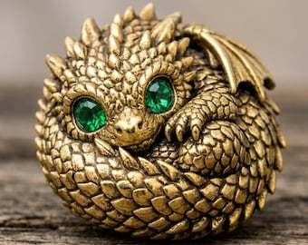 May include: A small, spherical brass figurine of a curled-up dragon with textured, scale-like detailing covering its body. The creature features two prominent, faceted emerald green gemstone eyes that stand out against the metallic gold finish.