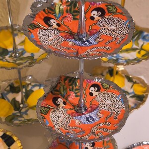May include: A tiered serving stand with decorative plates. The plates feature various designs, including a leopard print on an orange background, lemons, and swimmers. The stand has a silver and gold finish.