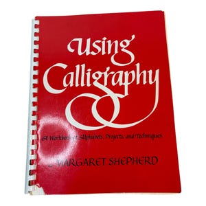 Könnte beinhalten: Ein rotes Arbeitsbuch mit dem Titel "Using Calligraphy" mit weißen Buchstaben. Das Buch hat eine Spiralbindung und enthält den Text "A Workbook of Alphabets, Projects, and Techniques" und den Namen des Autors, Margaret Shepherd.