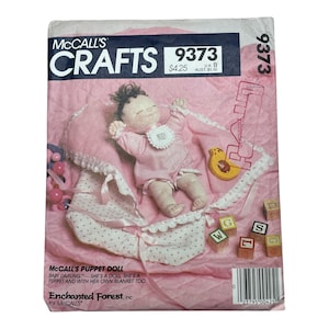 Könnte beinhalten: Eine McCall's Crafts 9373 Schnittmusterhülle mit einer Babypuppe in einem rosa gepunkteten Outfit und passender Decke. Das Cover zeigt den Text McCall's Crafts 9373, Enchanted Forest und McCall's Puppet Doll Baby Darling sowie einen Strichcode.