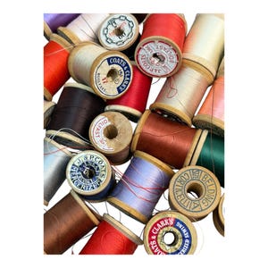 Puede incluir: Una colección de carretes de hilo de madera vintage en varios colores, incluyendo rojo, marrón y crema. Algunos carretes tienen etiquetas con textos como "Coats & Clark's" y "Boilfast". Los hilos están enrollados en los carretes, listos para coser.