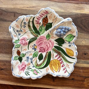 May include: A leaf-shaped ceramic serving dish with a white base, featuring a hand-painted floral design. The design includes pink roses, green leaves, and blue grapes. The rim is scalloped and outlined with blue paint.
