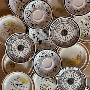 May include: A collection of ceramic plates and saucers featuring a mix of black and white floral patterns, geometric sunflower-like designs, and plates with yellow and brown botanical illustrations on a cream-colored background.