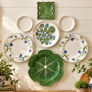 May include: A collection of decorative ceramic plates arranged on a wall. The set includes two white plates with blue floral patterns, one plate with a green and blue floral design, two small white scalloped plates, a square green plate, and a large green cabbage-shaped plate.