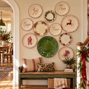 May include: A collection of eleven decorative ceramic plates arranged on a wall, featuring holiday themes like holly, poinsettias, a reindeer, Santa, and text reading Joy, Love, Hope, and Peace, above a wooden bench with two patterned pillows.