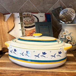 May include: A white ceramic casserole dish with a lid, featuring blue floral and leaf designs, and yellow and teal stripes. The dish has yellow handles and a yellow knob on the lid. It sits on a wooden cutting board.