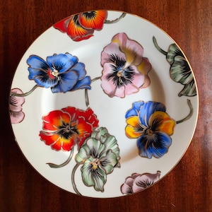 May include: A white ceramic plate with a gold rim, adorned with colorful pansy flower designs. The pansies feature shades of blue, pink, red, and green, creating a vibrant floral pattern. A decorative piece for serving or display.
