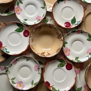 May include: A collection of ceramic dinnerware featuring multiple saucers and bowls with scalloped edges. The pieces are decorated with hand-painted pink flowers, green leaves, and dark red fruit motifs on cream and tan backgrounds with brown rims.