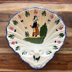 May include: A white, shell-shaped ceramic dish with a scalloped edge, hand-painted with a folk art design. The design features a figure in traditional clothing, flowers, and foliage. The rim is trimmed with blue paint.