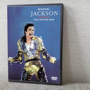 Puede incluir: Caja de DVD con Michael Jackson en el escenario, con una chaqueta y pantalones dorados y negros. El título "Michael Jackson" está en la parte superior, con "The Love We Save" debajo. El logotipo de DVD está en la esquina inferior derecha.