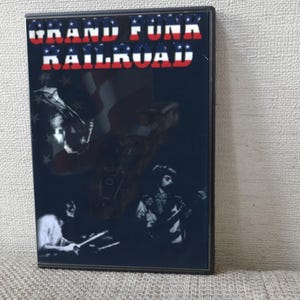 以下が含まれることがあります： グランド・ファンク・レイルロードのDVDケース。カバーには、バンド名が赤と白の文字で、青い背景にアメリカ国旗のデザインが施されています。バンドメンバーのシルエットが見えます。