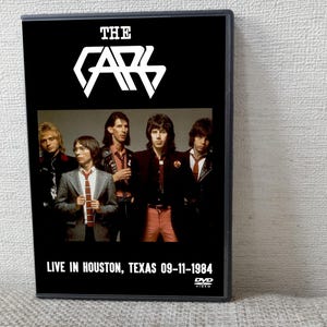 以下が含まれることがあります： 「The Cars Live in Houston, Texas 09-11-1984」のDVDケース。黒いケースにはバンドのロゴとメンバーの写真が写っています。DVDビデオケースはニュートラルな背景にあります。