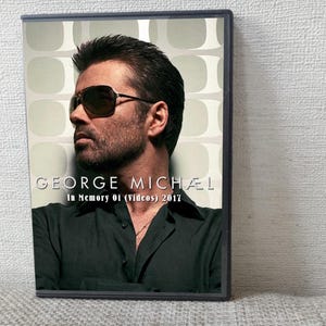 以下が含まれることがあります： サングラスをかけたジョージ・マイケルのDVDケース。テキストは「GEORGE MICHAEL In Memory 01 (Videos) 2017」です。背景には、薄緑色と白色の図形パターンがあります。ケースは黒いフレームに入っています。