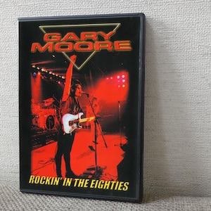 以下が含まれることがあります： ライブコンサートの画像が描かれたDVDケース。カバーには「GARY MOORE」の文字が赤と黄色で、下部には「ROCKIN' IN THE EIGHTIES」の文字が黄色で表示されています。画像には、赤い照明の中でステージに立つギタリストが描かれています。