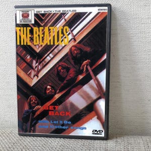 Könnte beinhalten: DVD-Hülle für "Get Back" von The Beatles. Das Cover zeigt die Bandmitglieder auf einem Balkon mit dem Bandnamen in Gelb. Die DVD-Hülle ist schwarz mit dem Wort "stereo" oben.