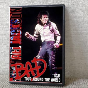 Puede incluir: Caja de DVD del concierto Bad Tour de Michael Jackson, mostrándolo en el escenario. La portada muestra "MICHAEL JACKSON" en rojo y azul, con "BAD" en letras rojas grandes. También se ve el texto "TOUR AROUND THE WORLD".