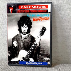 以下が含まれることがあります： ゲイリー・ムーアのDVDケース。赤と白で「GARY MOORE LIVE AT ISSTADION STOCKHOLM SWEDEN 1987」と書かれています。カバーアートは、ギターを持ったムーアの白黒画像です。「WILD FRONTIER」というタイトルは青色です。