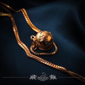 May include: A gold-colored necklace with a globe pendant, featuring continents and the word "Africa". The chain is a box chain style, and the pendant is suspended on a dark blue background. The necklace is likely made of gold or gold-plated metal.