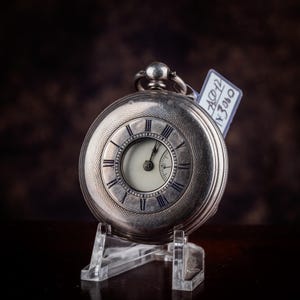 Könnte beinhalten: Eine silberne Taschenuhr mit weißem Zifferblatt und schwarzen römischen Ziffern. Die Uhr wird auf einem transparenten Ständer vor einem dunkelbraunen Hintergrund präsentiert. Ein Preisschild ist angebracht.