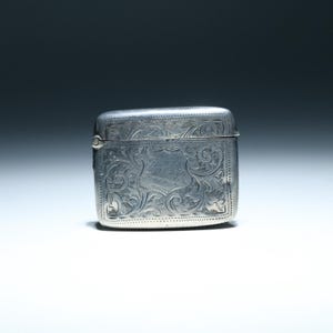 May include: A rectangular silver box with a hinged lid. The box is adorned with intricate floral and scrollwork engravings. The edges are subtly rounded, and the box has a small clasp. The overall design is ornate and detailed.
