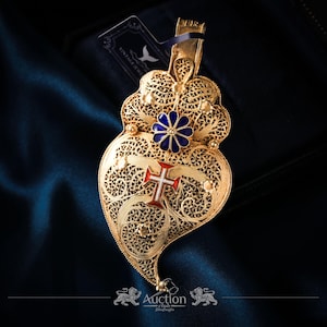 Könnte beinhalten: Ein goldener Filigrananhänger in Herzform mit einer blauen Blume und einem rot-weißen Kreuz. Der Anhänger hat aufwendige Durchbrucharbeiten und eine Schlaufe für eine Kette. Der Hintergrund ist ein dunkelblauer Stoff.
