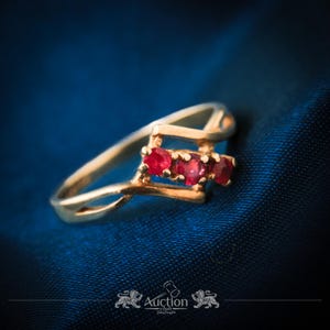 Op de afbeelding: Een gouden ring met drie rode edelstenen. De ring heeft een sierlijk, wervelend ontwerp en wordt gepresenteerd op een blauwe stoffen achtergrond. Een sieraad.