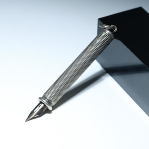 Könnte beinhalten: Ein silberfarbener Füllfederhalter mit einem strukturierten, rautenförmigen Schaft. Der Stift hat eine Feder und eine kleine Schlaufe oben. Der Stift ist gegen einen schwarzen Block auf einer weißen Oberfläche geneigt.