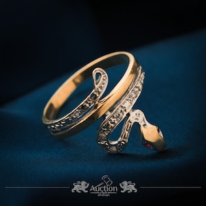 May include: A gold-tone snake ring with a diamond-studded band and a red gemstone eye. The ring is set against a dark blue background, showcasing the intricate design and craftsmanship.