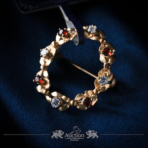 Puede incluir: Broche dorado con diseño de corona circular, adornado con piedras preciosas rojas y claras alternas. El broche tiene un cierre de alfiler y está sobre un fondo azul oscuro.