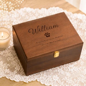 Könnte beinhalten: Eine rechteckige, dunkelbraune Erinnerungsbox aus Holz mit einem goldfarbenen Metallverschluss. Der Deckel ist mit dem Namen William, einem Pfotenabdruck-Symbol und dem Text graviert: In our hearts you are our dearest love. Our most difficult farewell.