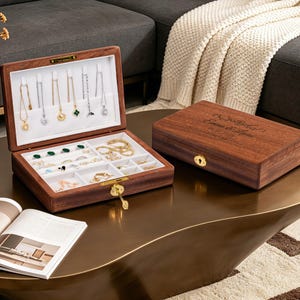 May include: Open wooden jewellery box with a white interior, displaying necklaces and rings. The box is brown with a gold clasp. A closed wooden box with engraved text is next to it. The jewellery box is on a gold-coloured table.