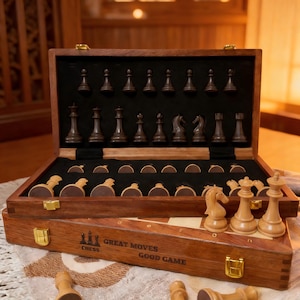 Op de afbeelding: Een houten schaakset met een donkerbruine afwerking, open om zwarte en lichtbruine schaakstukken te onthullen. De set bevat een houten kist met de tekst "Great Moves Good Game" en goudkleurige sluitingen. De schaakstukken zijn gerangschikt op een zwart vilt oppervlak.