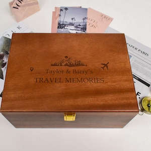 Puede incluir: Una caja de madera para recuerdos de viaje con el texto "Taylor & Barry's TRAVEL MEMORIES" grabado en la tapa. La caja tiene un cierre dorado y está rodeada de artículos temáticos de viaje, incluyendo postales y una brújula.