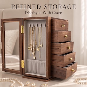 Puede incluir: Un joyero de madera oscura con puerta de cristal, cinco cajones extraíbles con pomos dorados y una sección interior para colgar collares con forro de tela gris. El texto REFINED STORAGE Displayed With Grace es visible en la imagen.