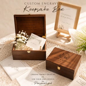 Puede incluir: Una caja de recuerdos de madera marrón oscuro con un cierre dorado. La tapa está grabada con las iniciales L M, los nombres LEWIS & MADELYN y la fecha MAY 04, 2023. La caja abierta contiene flores blancas de paniculata y una tarjeta.