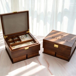 May include: Two wooden jewelry boxes. One is open, revealing compartments for rings, necklaces, and other jewelry. The other box is closed, with the word "MARORA" and a floral design engraved on the lid. Both boxes have gold-colored hardware.