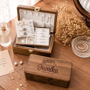 May include: A dark wood jewelry box with a hinged lid featuring the engraved name Jamika and the date 2026.04.26. The open box reveals a white velvet interior with compartments for earrings, necklaces, and rings, including several silver rings.