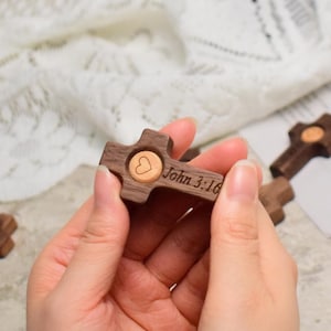 Op de afbeelding: Houten kruis met een hartvormige inleg en de inscriptie "Johannes 3:16". Het kruis is donkerbruin met een lichter houten hart. Er zijn meerdere kruisen zichtbaar.