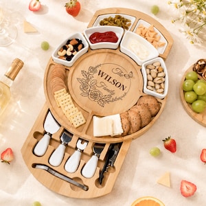 May include: A round bamboo charcuterie board with a slide-out drawer containing four cheese knives and a corkscrew. The centre is engraved with "The WILSON Family" surrounded by roses and a butterfly. It holds white ceramic bowls and snacks.