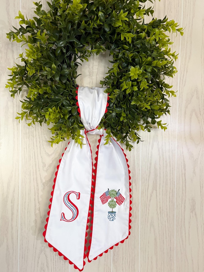 May include: A green wreath with a white and red decorative scarf. The scarf has a red scalloped edge and features an embroidered letter "S" and a design of American flags. The wreath is hanging on a light wood-panelled wall.