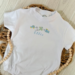 May include: A white short-sleeved baby romper featuring an embroidered design of a blue car, a green airplane, and a blue train connected by a black stitched line. Below the vehicles, the name Ellis is embroidered in light blue cursive script.