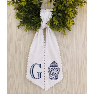 May include: White decorative towel with a scalloped edge, embroidered with a blue letter "G" and a blue and white ginger jar design. The towel is hanging from a green wreath.